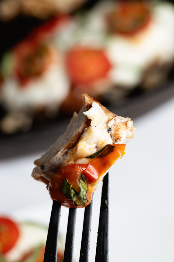 Freshly prepared Grilled Chicken Caprese with Balsamic reduction on a fork in front of a cast iron grill pan with another prepared chicken breast. 