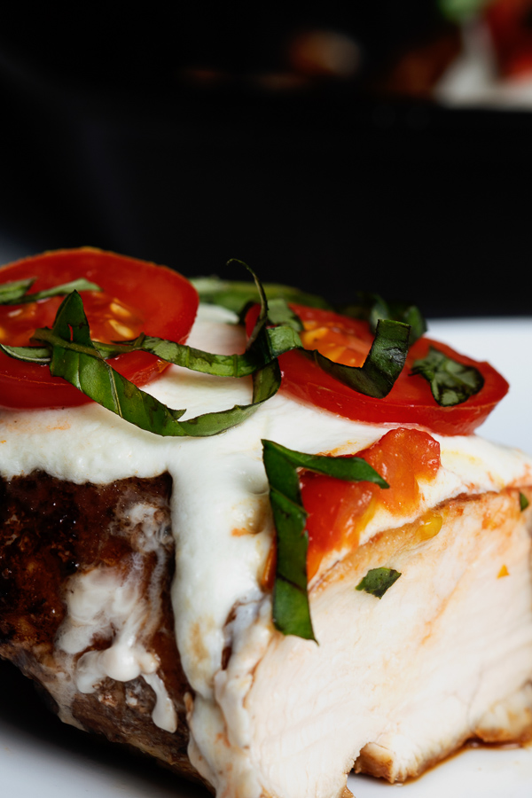 A close-up view of fresh basil and small red tomatoes atop a bed of melted Mozzarella cheese, covering a balsamic glazed chicken breast. 