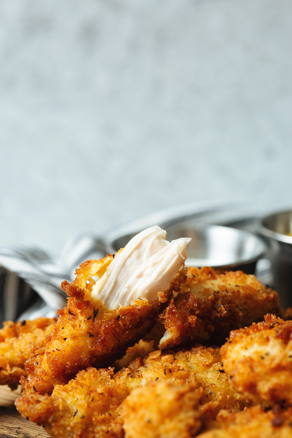 A broken fried chicken tender in the foreground on top of a pile of whole fried chicken tenders, shows the white moist meet inside. In the background a white and blue napkin with two silver ramekins of dipping sauces. 