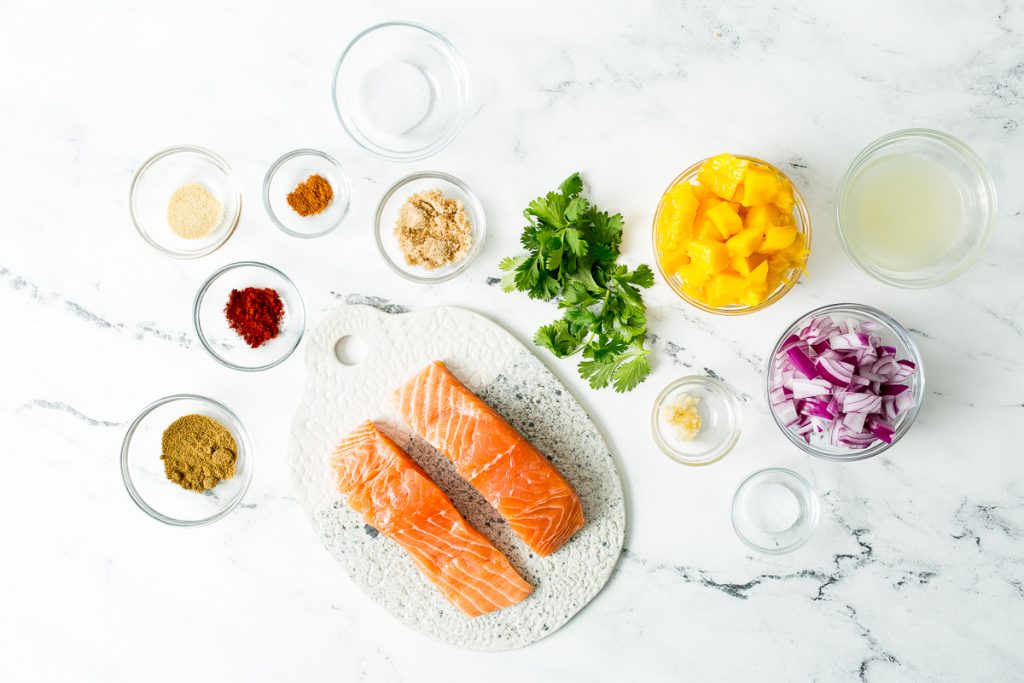 Overhead image of the ingredients required for blackened salmon with mango salsa: two salmon filets, salt, brown sugar, cayenne, onion powder, smoked paprika, cumin, cilantro, mango, purple onion, lime juice, garlic, and salt. All in glass bowls on a white marble countertop. 