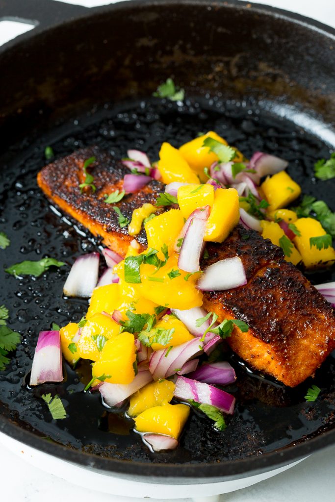 A close up image of blackened salmon topped with mango salsa in a cast iron skillet. 