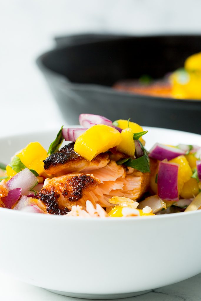 Close up image of blackened salmon topped with mango salsa in a white bowl with rice. In the background a cast iron skillet with blackened salmon and mango salsa.