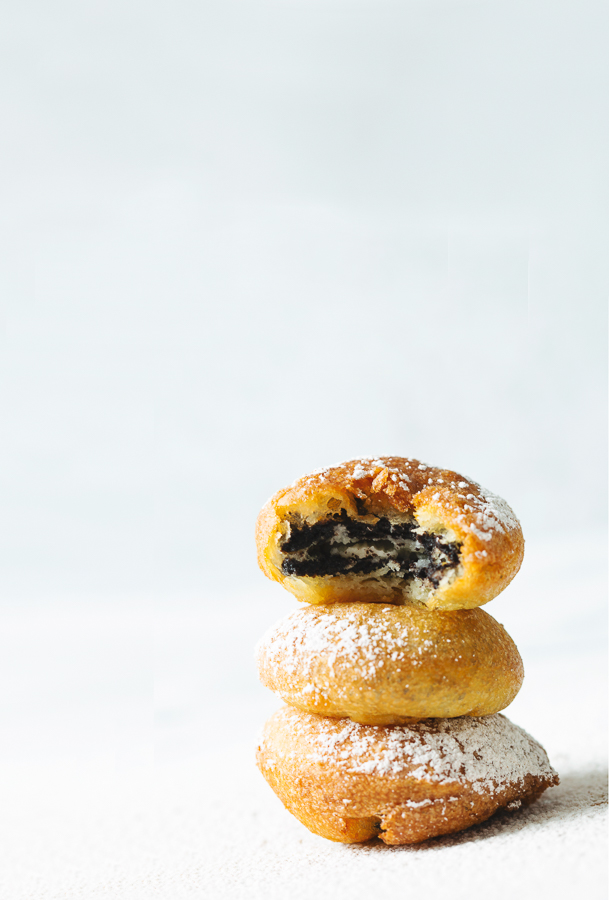 Three homemade deep fried oreos dusted with cinnamon powdered sugar are stacked on top of each other with a white background. The top deep fried oreo has a single bite taken out of it, exposing the inside of this favorite fair food. 