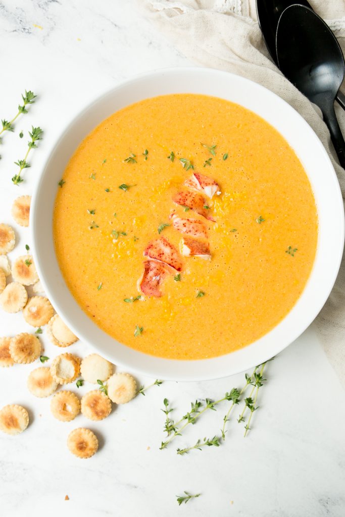 A clean white bowl full of bright orange lobster bisque, topped with more fresh lobster meat and fresh thyme, sits on a white marble table. The bowl is surrounded by oyster crackers, fresh thyme, two black spoons and an antique style tan cloth napkin.