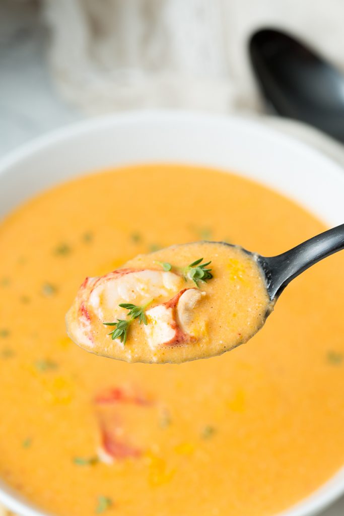 A black spoonful of lobster bisque topped with fresh lobster meat and fresh thyme in the foreground hovers over a white bowl of lobster bisque in the background. 