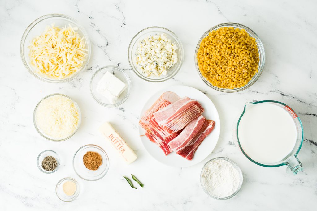 Image: An overhead view of the ingredients for this Dutch Oven Mac and Cheese individually placed in glass bowls and a white plate. The ingredients include: Cheddar cheese, blue cheese, gruyere cheese, cream cheese, elbow macaroni, bacon, butter, fresh sage, pepper, nutmeg, garlic powder, flour, and whole milk.