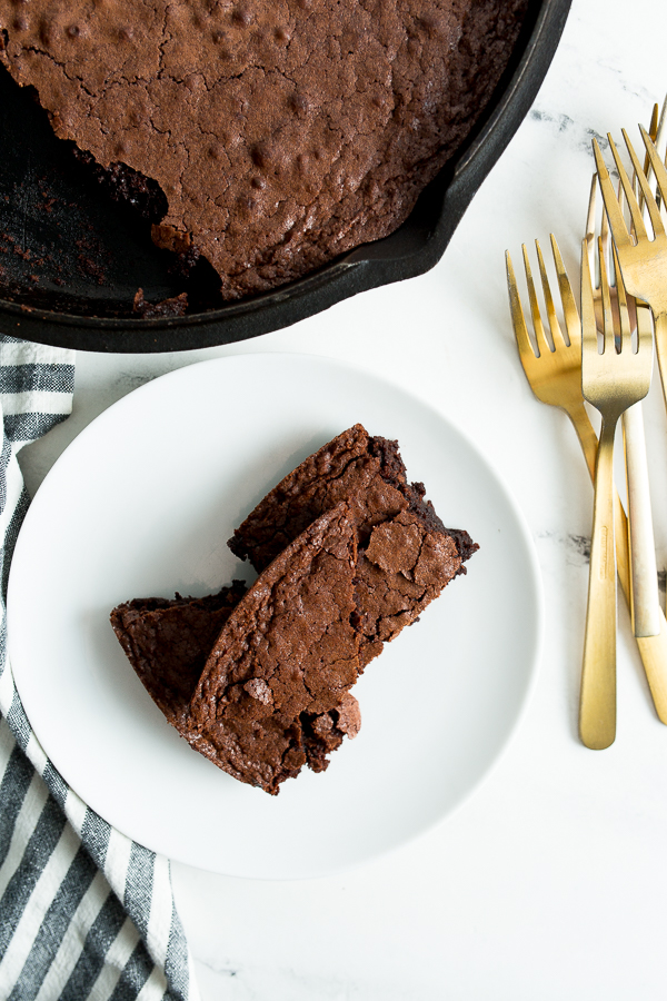 Overhead image of chocolate brownies on a white plate surrounded by gold forks, blue and white cloth napkin, and a cast iron skillet. 