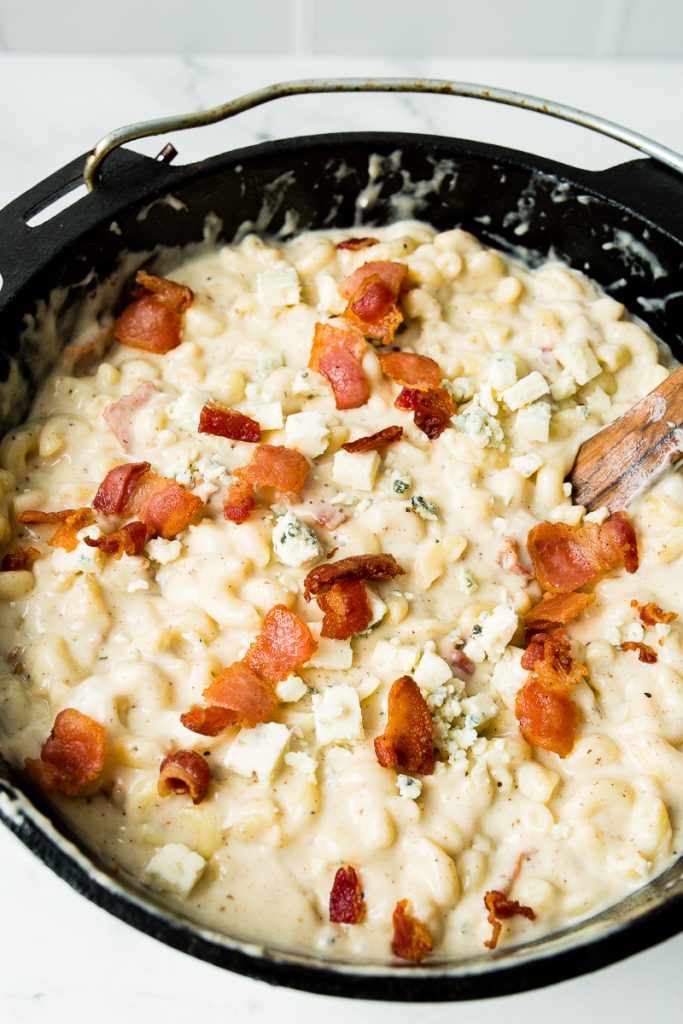 Image: an top view of a dutch oven filled with mac and cheese, topped with bacon and fresh blue cheese. 