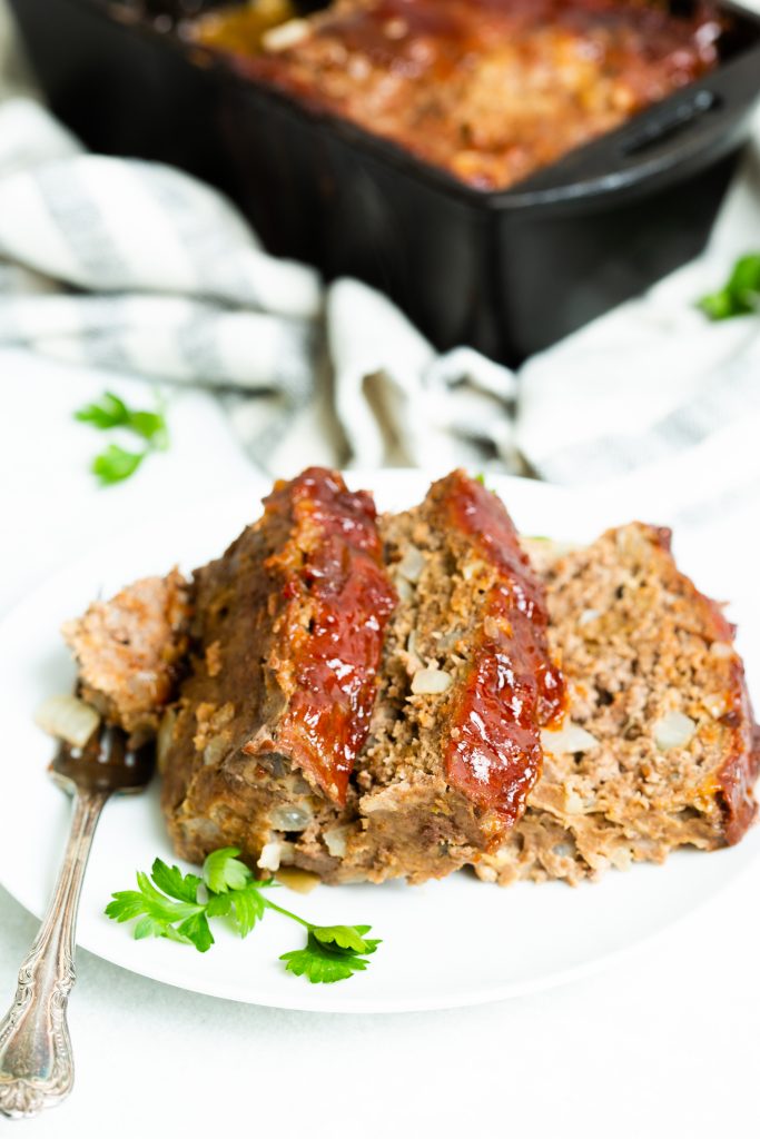 Image of: three slices of meatloaf with ketchup glaze on a white plate and a cast iron loaf pan in the background. 