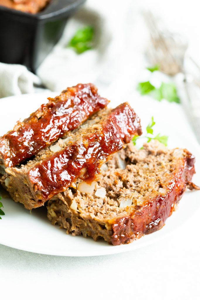 Image of: three slices of meatloaf on a white plate and cast iron loaf pan in the background. 