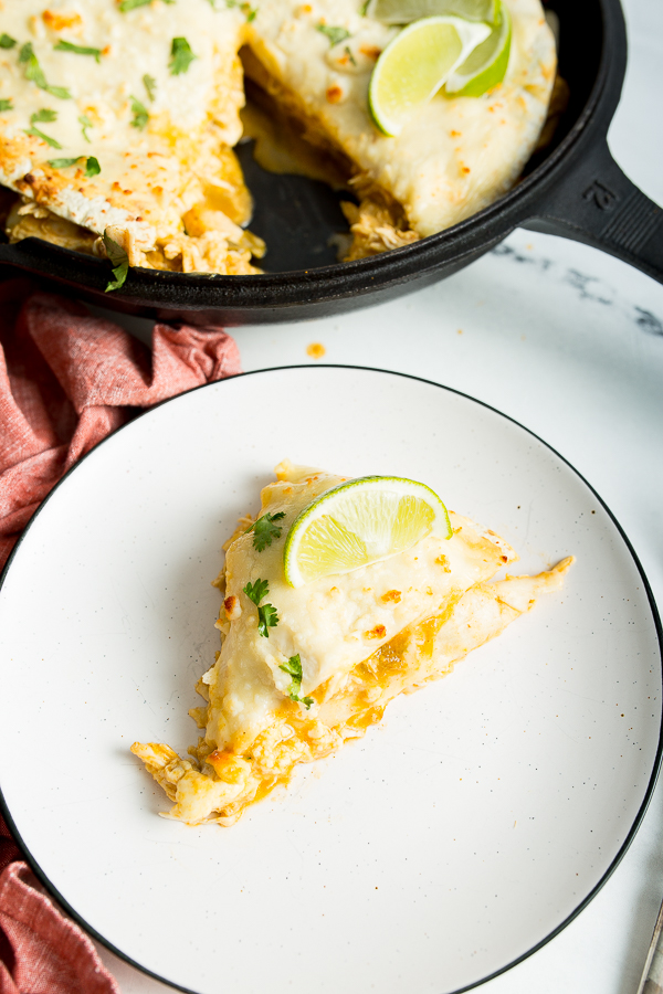 A close up image of green enchilada lasagna on a plate topped with cilantro and lime wedge, with a cast iron skillet in the background.