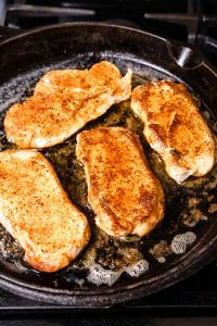 A side process shot of four boneless pork chops in a cast iron skillet on the stove.