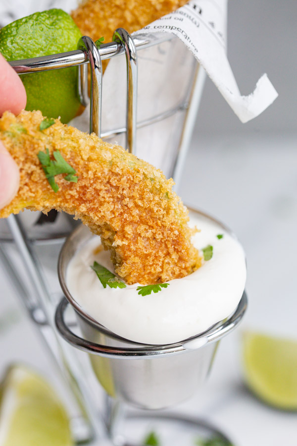 A side view of an avocado fry being dipped into a small dish of lime crema. 