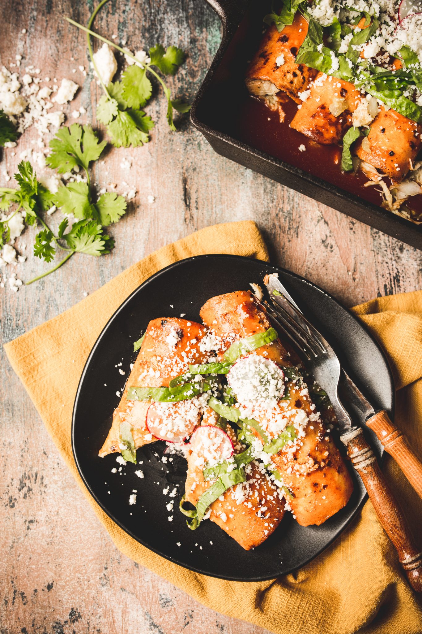 Overhead image of red chicken enchiladas on a black serving plate topped with radishes, lettuce, and cotija cheese. 