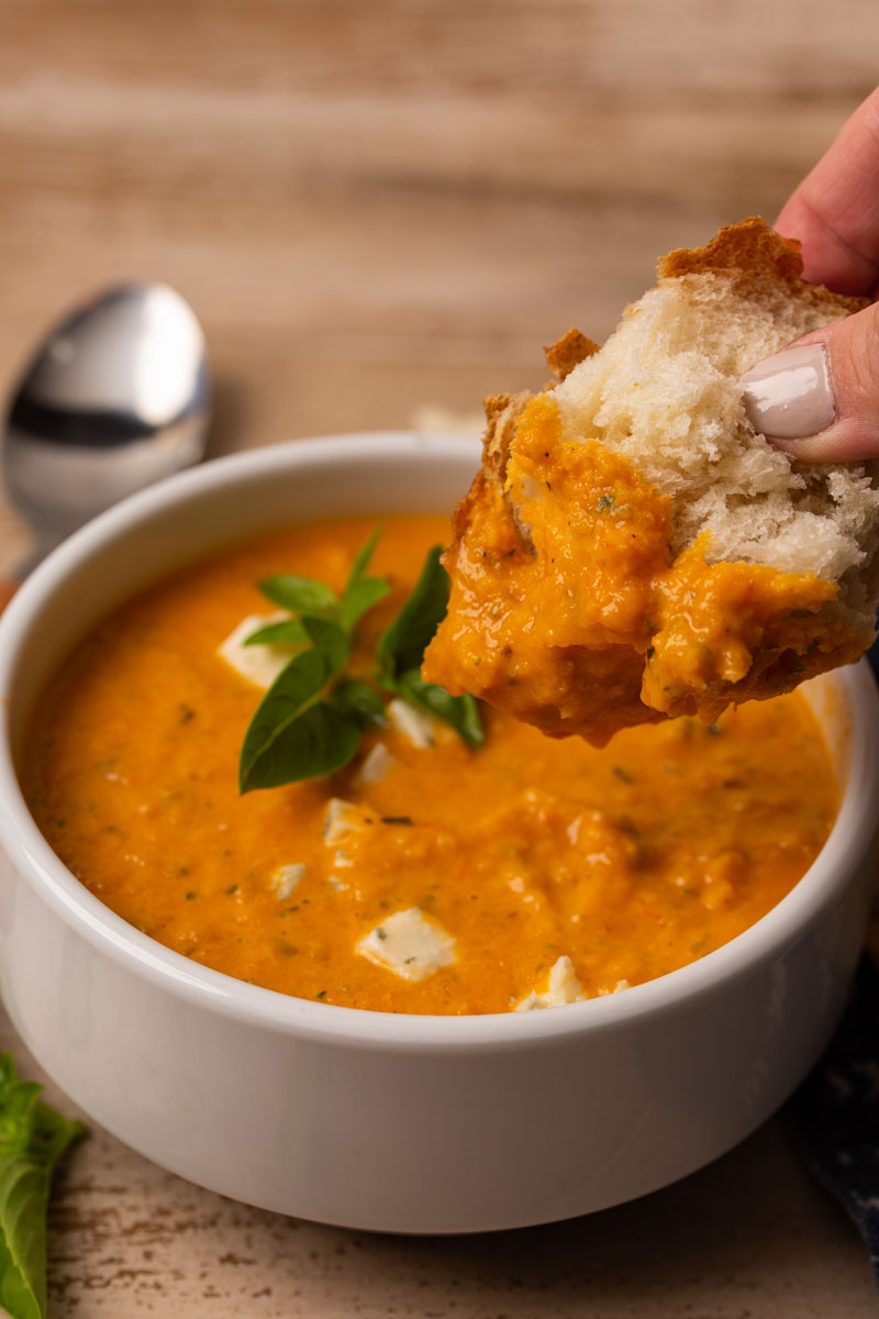Close up image of tomato basil soup with bread being dipped in and fresh basil garnished on top. 