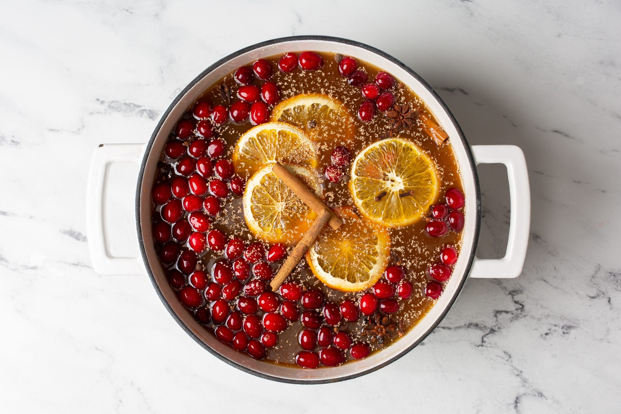 Overhead process image of mulled apple cider being made in a dutch oven with oranges, cranberries, cinnamon and spices.