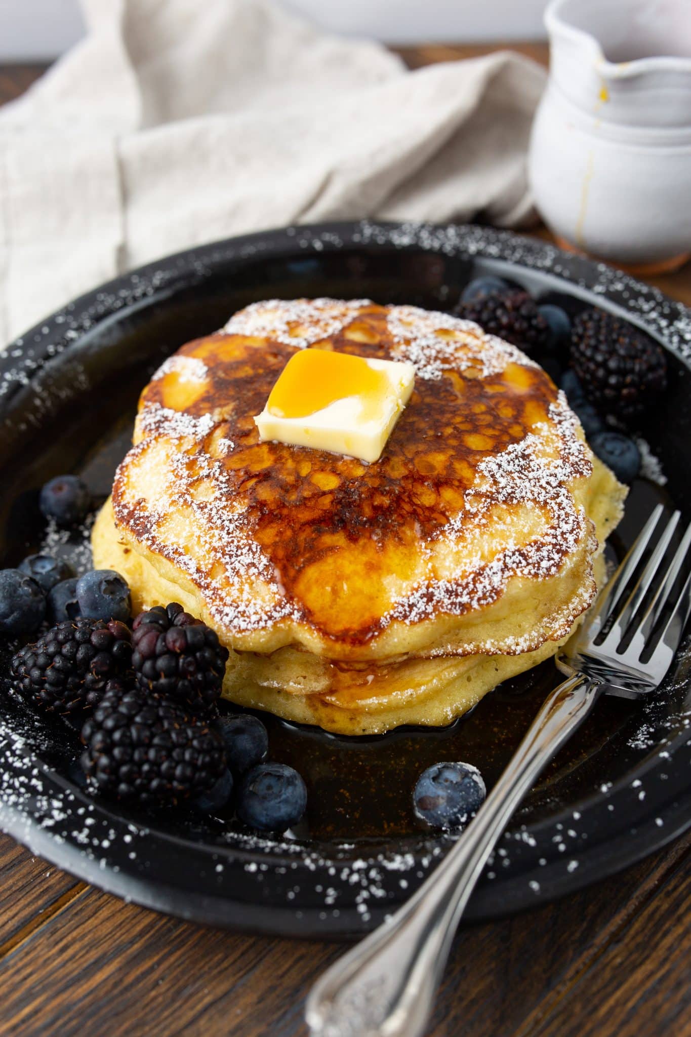 stack of buttermilk pancakes on a plate with pad of butter, maple syrup, powdered sugar, and fresh berries. 