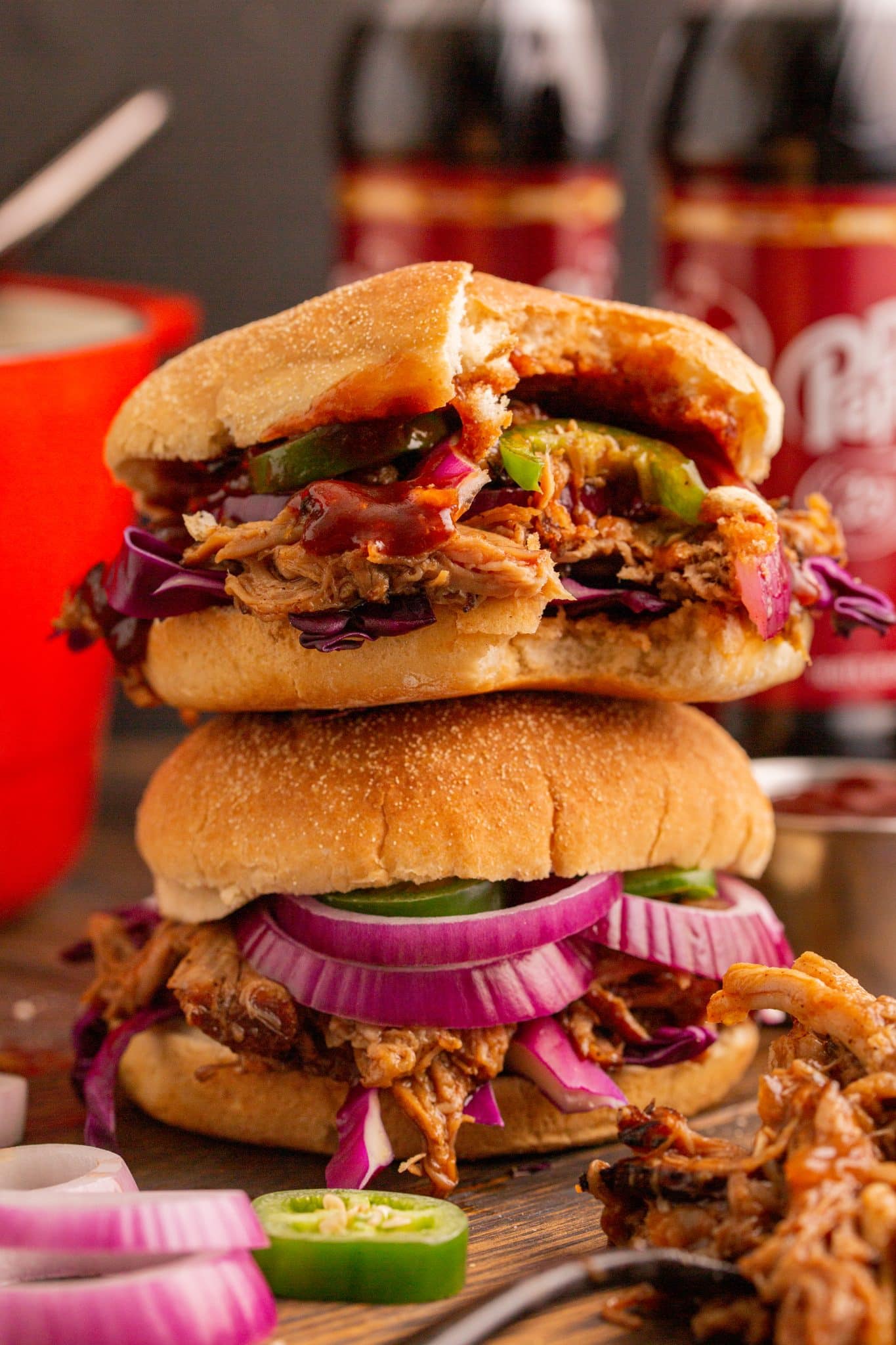 Two Dr Pepper Pulled Pork sandwhiches stacked on top of each other dripping with bbq sauce. Dr pepper and dutch oven in the background. 