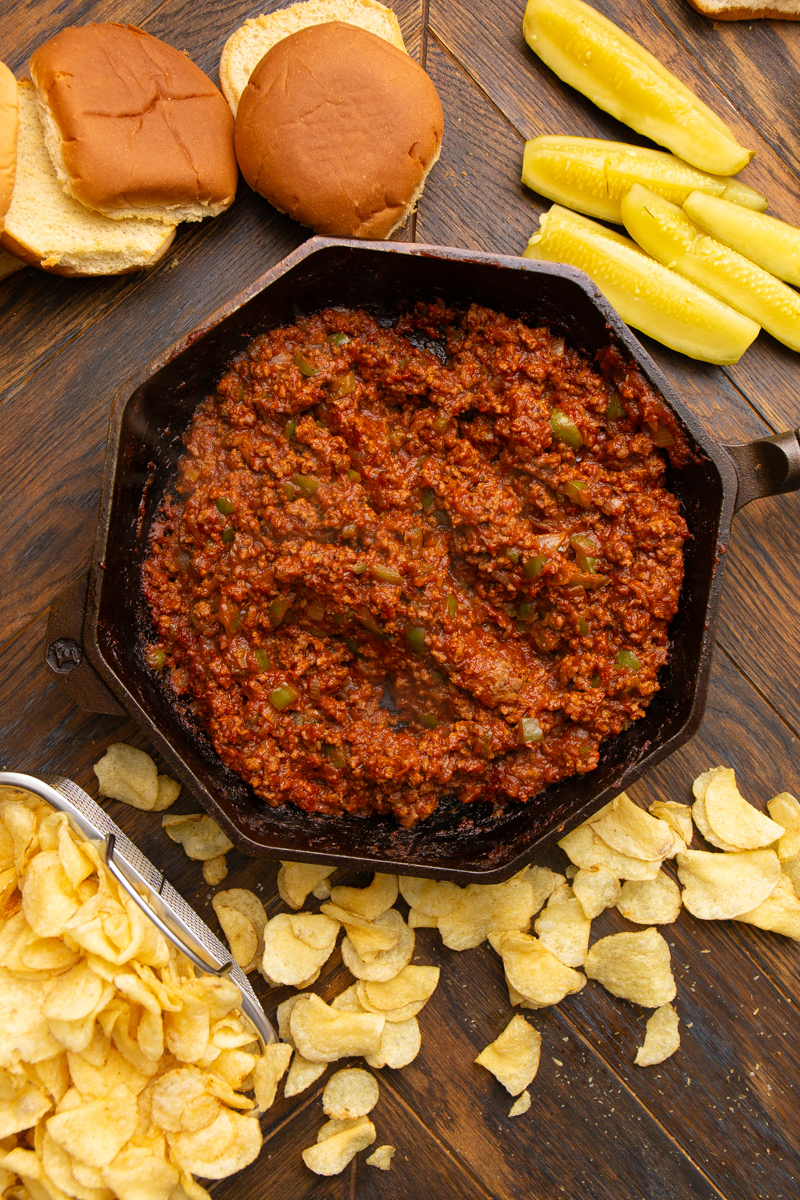 overhead image of sloppy joes in a skillet surrounded by buns, pickles, and chips
